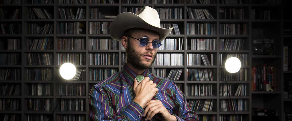 Charley Crockett wearing a striped shirt, cowboy hat and adjusting his tie.